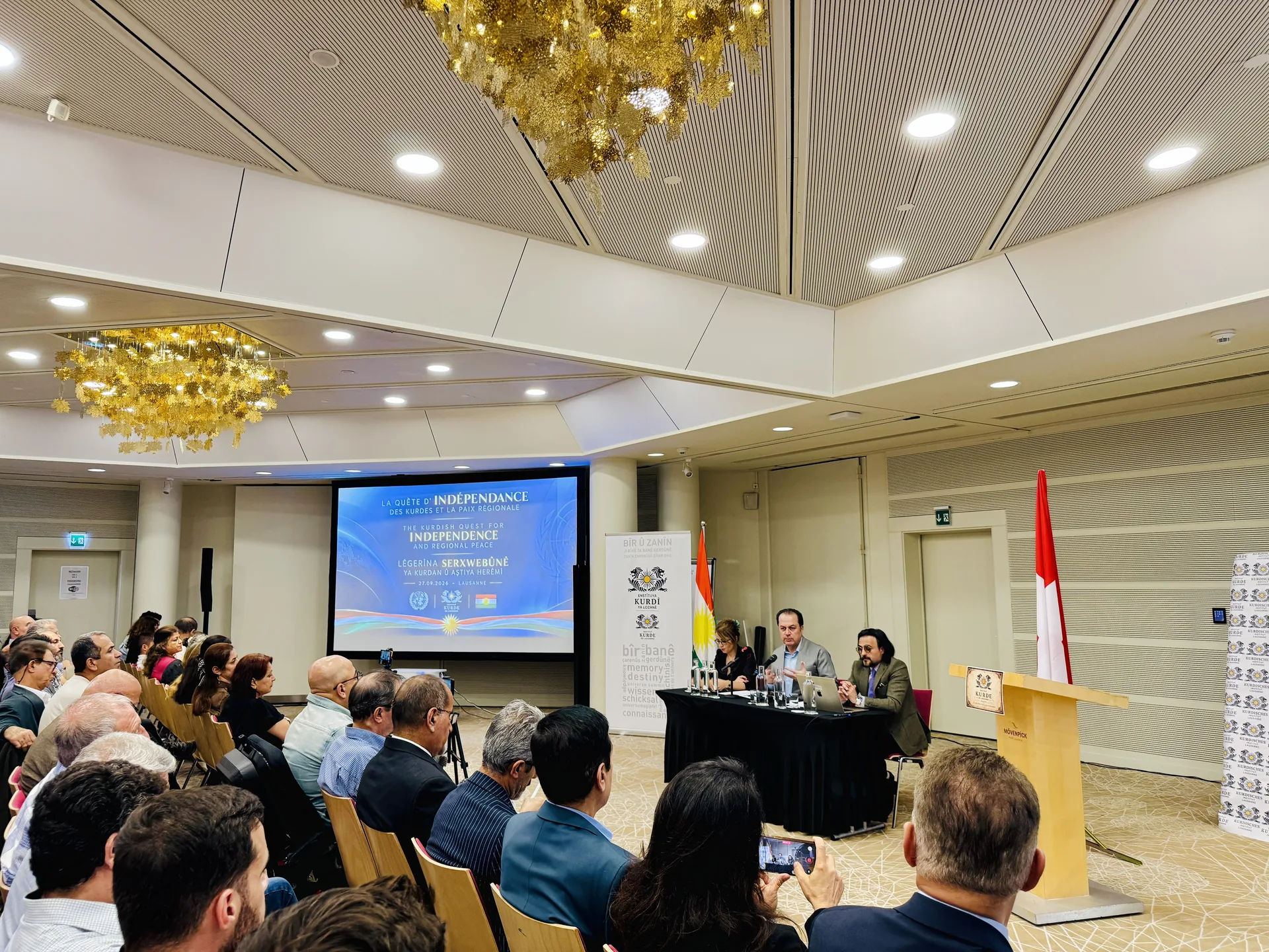 Salona konferansê ya Mövenpick Hotelê: beşdar, maseya axavtvanan û ekrana konferansê, Lozan, 27.09.2026